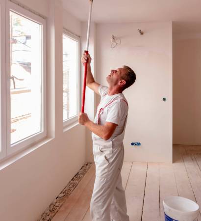 Construction worker painter man with paint roller in his hand and buckets of products to restore and paint the wall and celling, indoor the building site of a house